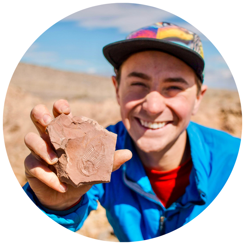 Headshot of Jeremy Snyder, a white man in his twenties, grinning while holding a trilobite fossil